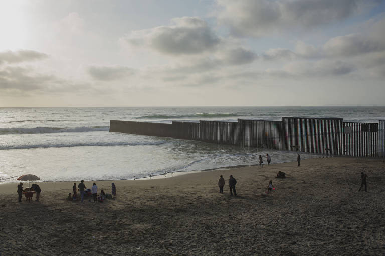 um praia, pessoas na praia e, partindo da areia e avanÃ§ando ao mar, um muro